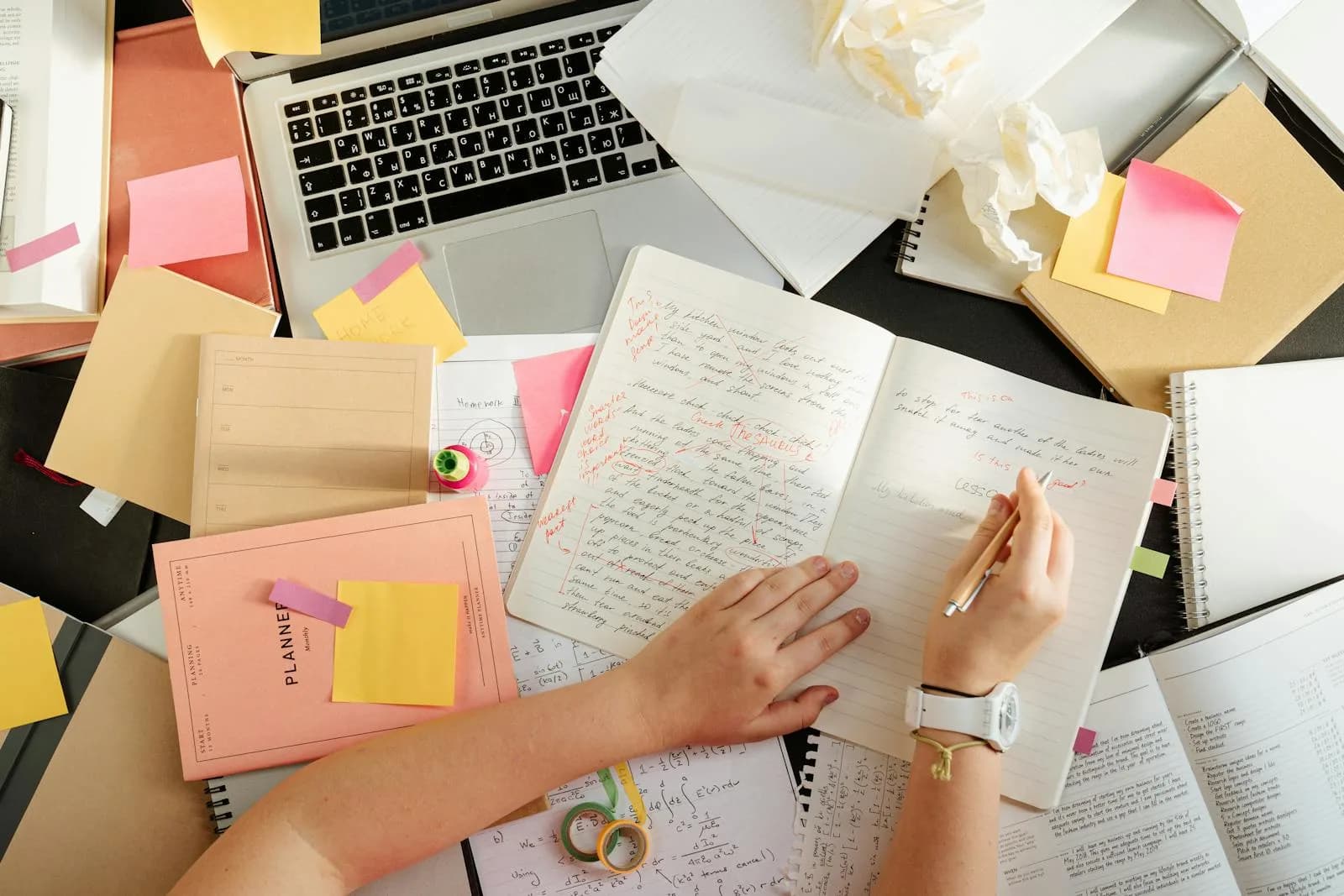 Plan aérien d'une personne organisant des notes et du matériel d'étude sur un bureau encombré avec un ordinateur portable.